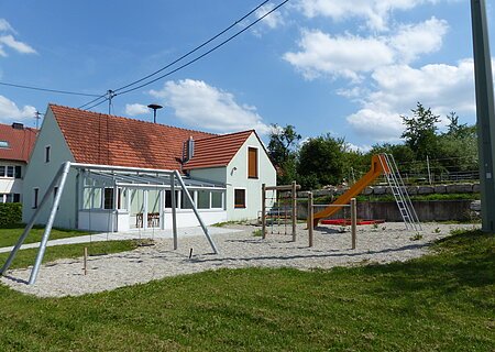 Spielplatz Liederberg Spielplatz mit einer Schaukel, Klettergerüst, einem Sandkasten mit roter Umrandung und oranger Rutsche, vor einem türkisfarbenen Haus mit rotem Dach, bei blauem Himmel. Umgeben von einer grünen Wiese und Bäumen mit Kieselsteine an den Spielgeräten.