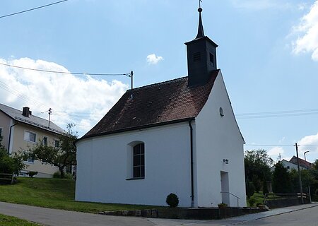 Filialkirche St. Johannes Nepomuk Liederberg Kleine weiße Filialkirche mit rotem Ziegeldach und kleinem Turm oben drauf mit Kreuz an einer Straße bei klarem Himmel. Dahinter ein weiteres Wohnhaus mit grüner Wiese und Bäumen.