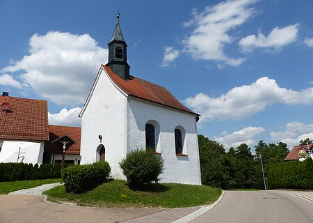 Filialkirche St. Kastulus Ried Kleine weiße Filialkirche mit rotem Dach und kleinem Turm oben drauf an einer Straße bei blauem Himmel mit Wolken. Umgeben von grüner Wiese und Hecken, daneben ein Jesuskreuz aus Holz. Im Hintergrund weiteres weißes Haus zu sehen.