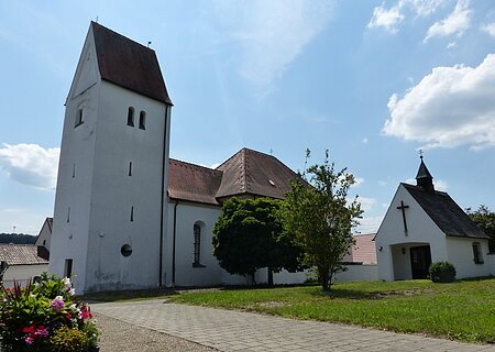 Filialkirche St. Nikolaus Warching Filialkirche mit rotem Dach und einer kleiner Kapelle daneben mit einem Turm bei blauem Himmel und grünem Rasen im Vordergrund. Neben dem Rasen verläuft ein gepflasterter Gehweg mit Schottersteinen daneben.