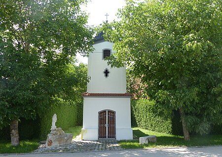 Marienkapelle Rothenberg Kleine weiße Marienkapelle mit Fenster und einem Eingang, umgeben von Bäumen und Grünflächen an einer Straße. Vor der Kapelle links ein kleiner Brunnen aus Stein und ein großer Stein rechts als Sitzgelegenheit.