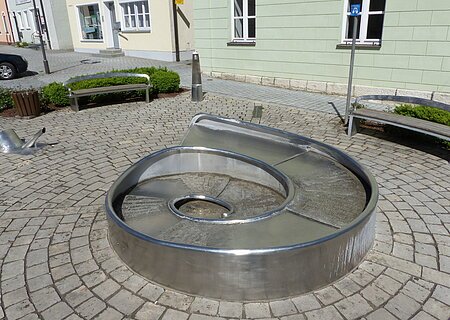 Juraschneck-Brunnen Monheim Juraschneck-Brunnen: ein Brunnen aus Metall in Form eines Fossils einer Schnecke, mit eingravierten Texten auf der Oberfläche. Außenrum ein gepflastertem Platz mit zwei Sitzbänken. Im Hintergrund farbige Häuser und eine kleine grüne Hecke an den Bänken entlang.