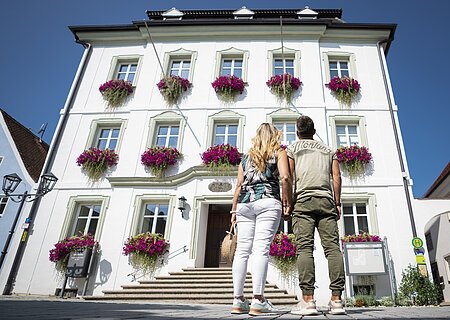 Das Rathaus in Monheim Paar, Mann und Frau von hinten vor einem weißem Gebäude mit Blumen in Blumenkästen an den Fenstern, schauen auf das Gebäude bei klarem Himmel. Eine Infovitrine auf der rechten Seite am Gebäude.