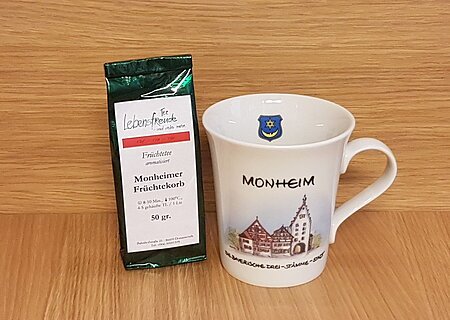 Neue Stadt Monheim-Tasse Geschenkidee der Stadt Monheim: eine weiße Tasse mit Monheim-Motiv und der Aufschrift "Monheim - Die Drei-Stämme-Stadt" rechts im Bild. Links davon eine Teeverpackung mit der Mischung und dem Text „Monheimer Früchtekorb“ stehen vor einer Holzoberfläche.