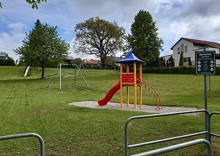 Spielplatz Monheim Bergstraße Spielplatz mit Rutsche, Klettergerüst, Seilbahn, Schaukel und Hinweisschild auf einer Wiese vor Wohnhäusern bei bewölktem Himmel.