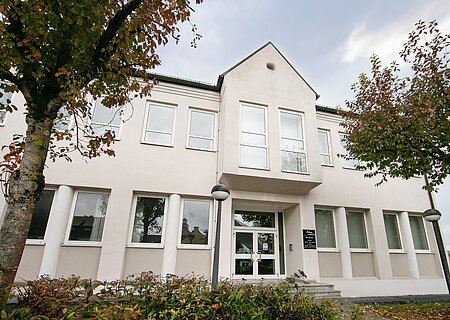 Appl Bürogebäude, Jahnstraße 19, 86653 Monheim Außenansicht Zweistöckiges Bürogebäude mit heller Fassade, mehreren Fenstern, zweiflügeliger Eingangstür aus Glas, Bäume und Büsche im Vordergrund, bei bewölktem Himmel im Hintergrund.