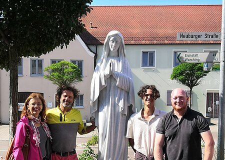 Marienstatue "Maria, Königin des Friedens" Vier Personen stehen neben einer weißen Marienstatue im Blumenbeet an einer Straße mit Häusern im Hintergrund.