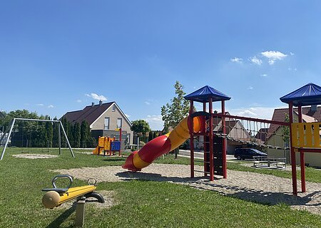 Spielplatz Monheim Am Krautgarten Spielplatz mit Wippe, Rutsche, Klettergerüst, Schaukel und Karussell bei sonnigem Himmel in einem Wohngebiet.