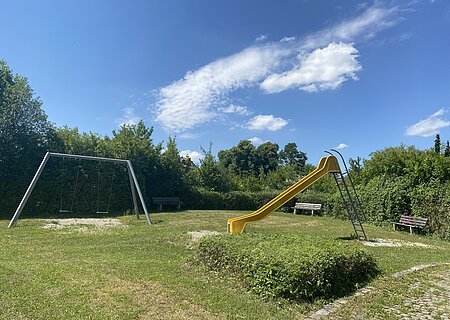 Spielplatz Finkenweg, Rotbrunnstraße Spielplatz mit gelber Rutsche, zwei Schaukeln und drei Bänken, umgeben von Büschen unter blauem Himmel.