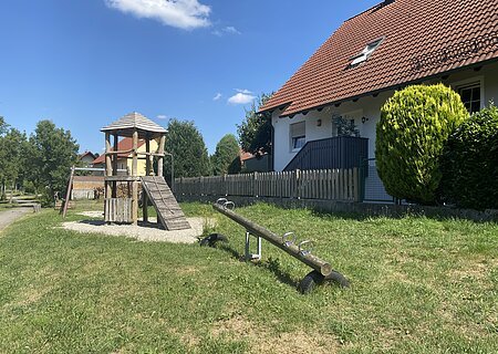 Spielplatz Kastanienweg Spielplatz mit Wippe, Schaukel im Hintergrund und Kletterturm auf grünem Rasen, links vor einem Haus mit rotem Dach und Zaun an einem sonnigen Tag.