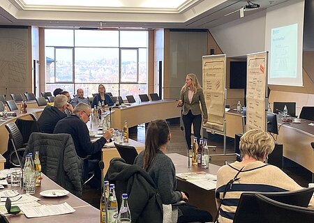 Pressebild - Austauschtreffen - Flächensparen Seminarraum mit hellen Tischen und schwarzen Stühlen, mehreren Personen sitzen an den Tischen, eine Frau präsentiert vor Flipcharts und einem Projektorbild.