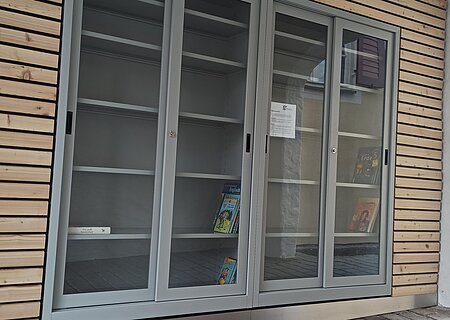 Offener Bücherschrank Monheim Außenansicht eines Bücherschranks mit Glastüren, teilweise mit wenigen Büchern, an einer Putzwand.