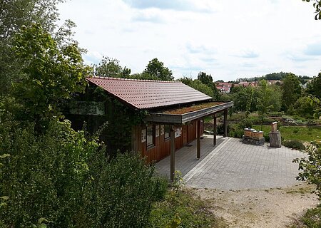 Lehrbienenstand mit Bienenweide Monheim Holzgebäude mit rotem Dach und begrüntem Vordach, umgeben von Bäumen und einem gepflasterten Vorplatz, bei bewölktem Himmel.