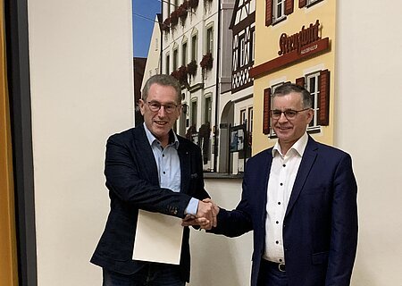 Kommunale Dankurkunde an Norbert Meyer Zwei Männer in Anzügen schütteln sich die Hand vor einer Wand mit einem Bild von Fachwerkhäusern. Der Mann links hält eine Urkunde in der anderen Hand.