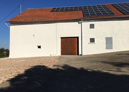 Ehemaliger Getränkemarkt Weilheim - Außenansicht Weißes Gebäude mit braunem Tor, drei kleinen Fenstern und Solarpaneelen auf rotem Ziegeldach bei klarem Himmel. Im Vordergrund geteerter Platz.