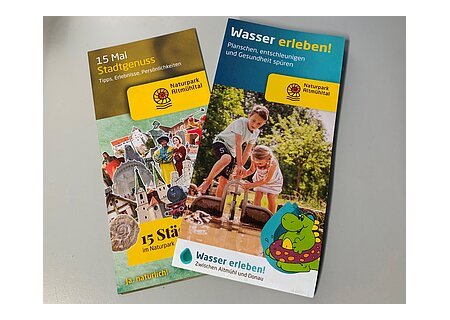 Naturpark "Altmühltal Wasser erleben & 15 Mal Stadtgenuss" Zwei Broschüren des Naturparks Altmühltal zu Stadtgenuss und Wassererlebnissen mit Bildern und Texten liegen übereinander auf einem weißen Tisch.
