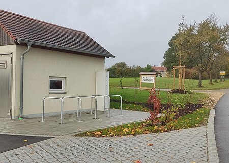 E-Bike Ladestation Flotzheim Kleines Gebäude mit Metalltür und kleinem Fenster, sowie fünf Fahrradständern auf gepflastertem Platz neben einer Straße und einem Schild mit Ortsnamen Flotzheim, unter bewölktem Himmel.