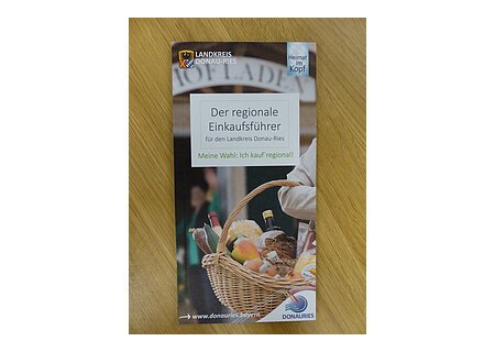 Der regionale Einkaufsführer 2019 Auf einem Holztisch die Broschüre des regionalen Einkaufsführers aus dem Landkreis Donau-Ries vom Herausgeber Ferienland Donau-Ries und deren zwei Logos auf dem Cover. Auf dem Foto ein Oberarm einer Person, die ein Flechtkorb voller Lebensmittel trägt mit unscharfen Hintergrund. Darüber mittig der Titel "Der regionale Einkaufsführer".