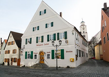 Metzger by Ferber Graues mehrstöckiges Gebäude mit grünen Fensterläden, einer Treppe mit Geländern und dem Schriftzug "Metzgerei Ferber" oberhalb der Eingangstür, an einer gepflasterten Straße. Links von der Tür der Schriftzug "Metzgerei", rechts davon "Imbiss" und "Pension" an der Fassade. Ein gelber Briefkasten hängt an der vorderen Fassade.