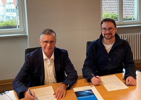 Vertragsunterzeichnung am 14.10.2024 mit Fa. Felkatec Zwei Männer blickend in die Kamera sitzen an einem Holztisch und unterschreiben Dokumente. Auf dem Tisch liegt eine Mappe mit der Aufschrift „GIGABIT FÖRDERBESCHEID“.