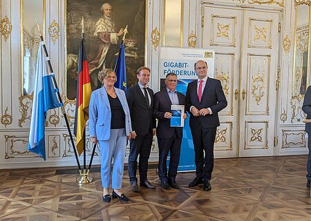 Übergabe des Zuwendungsbescheids am 11.10.2024 im Rokokosaal in Augsburg Fünf Personen in Anzügen stehen vor drei Fahnen und einem Banner mit der Aufschrift „Gigabit-Förderung“ in einem prunkvollen Raum. Rechts im Bild steht ein weiterer Mann im Anzug am Rednerpult.