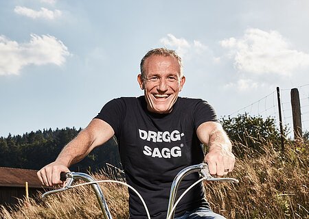 Michl Müller "Limbo of Life" Mann mit schwarzem T-Shirt mit der weißen Aufschrift "DREGG-SAGG" und Jeans sitzt lachend auf orangefarbenem Fahrrad auf Feldweg bei Sonnenschein