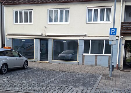 Mietobjekt Neuburger Straße 9 - EG Außenansicht Zweistöckiges zweifarbiges Gebäude mit drei Fenstern oben und drei großen Schaufenstern unten. Ein parkendes Auto auf einem Parkplatz davor.