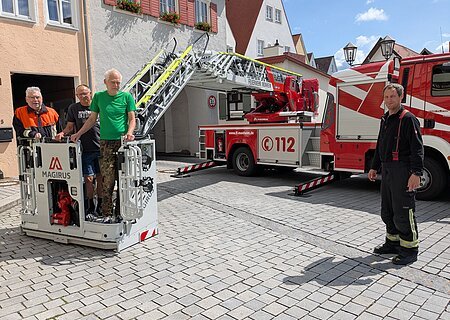Das "Storchenteam" bei der Storchenberingung 2025 Drei Männer in ziviler Kleidung stehen in einem Feuerwehrkorb vor Wohnhäusern und Torbogen, ein Mann in Feuerwehr-Uniform steht rechts daneben, im Hintergrund ein Feuerwehrfahrzeug mit ausgefahrener Drehleiter, bei blauem Himmel.