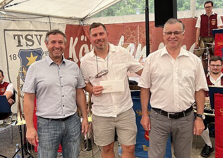 TSV Monheim Spendenübergabe Drei Männer stehen nebeneinander vor einem Zelt mit Musikern im Hintergrund, einer hält einen Briefumschlag. Hinter ihnen ein Banner mit Aufschrift TSV 1895 Monheim sichtbar.