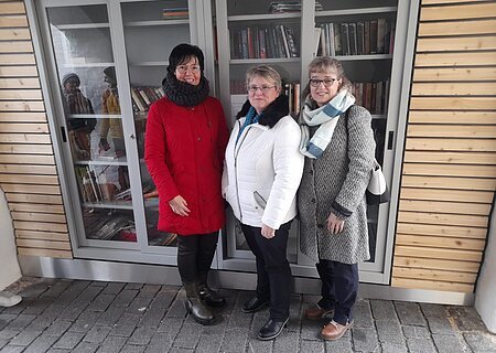 Offizielle Eröffnung des offenen Bücherschrankes Drei Frauen in Winterkleidung stehen vor einem Bücherschrank mit Glastüren links und rechts am Schrank eine Holzfassade.