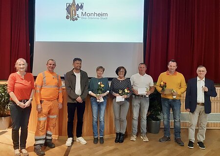Personalrat - Ehrungen 2024 Acht Personen stehen nebeneinander vor einer Bühne mit rotem Vorhang und einer Leinwand mit Monheim-Logo, einige halten Blumen und Urkunden.