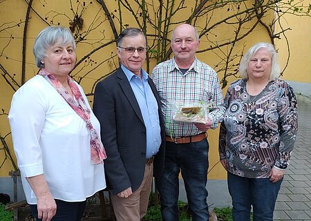 Schulverband Ehrung und Verabschiedung Vier Personen stehen nebeneinander vor einer gelben Wand mit kahlen Ästen, eine Person hält ein verpacktes Geschenk.