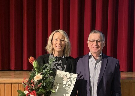 Dienstjubiläum Evelyn Wagenknecht Zwei Personen vor rotem Bühnenvorhang, eine hält Blumenstrauß und Urkunde, beide lächeln in die Kamera blickend.