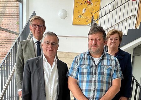 Ehrung Feldgeschworene Vier Personen stehen blickend in die Kamera auf einer Treppe in einem Gebäude mit schwarzem Geländer und großem Fenster. An der Wand hängt ein gelbes Gemälde.