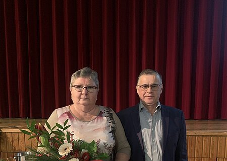 Verabschiedung Frau Brunhilde Biller Zwei Personen vor rotem Bühnenvorhang, Frau hält Blumenstrauß, Mann trägt Anzug und Brille. Im Hintergrund ein gedeckter Tisch.