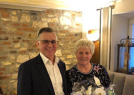 Verabschiedung Lisa Wiedemann Zwei Personen, ein Mann und eine Frau stehen in einem Raum mit Steinwand im Hintergrund und einem Sitzsofe. Vor der Frau steht eine verpackte weiße Orchideenpflanze auf einem Holztisch.