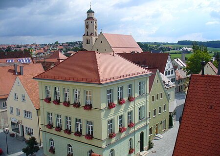 Haus des Gastes - im Hintergrund die Stadtpfarrkirche Monheim Blick von oben auf eine Innenstadt mit Hauptaugenmerk auf ein historisches grünes Gebäude im Vordergrund, mit roten Dachziegeln und Blumen an den Fenstern. Im Hintergrund weitere Häuser der Kleinstadt und blauer Himmel. Rechts unten im Eck das rote Dach eines anderen Gebäudes im Vordergrund zu sehen.