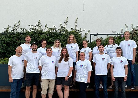 Das Team der Wasserwacht Monheim Gruppenfoto von 15 Personen in weißen T-Shirts mit dem Logo der Wasserwacht drauf, stehen blickend in die Kamera vor einer weißen Wand mit Pflanzen im Hintergrund.