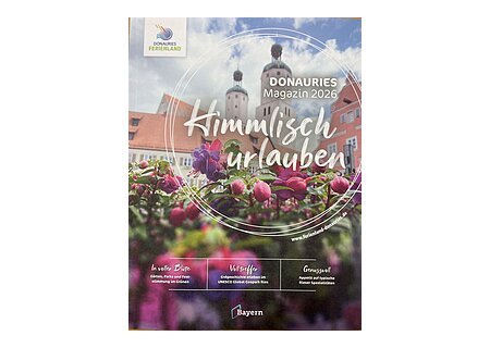 DONAURIES Magazin 2026 "Himmisch urlauben" Blumen im Vordergrund, dahinter Gebäude mit zwei Türmen, Bayern-Logo, Text zu Donauries Ferienland Magazin 2026 "Himmlisch urlauben"