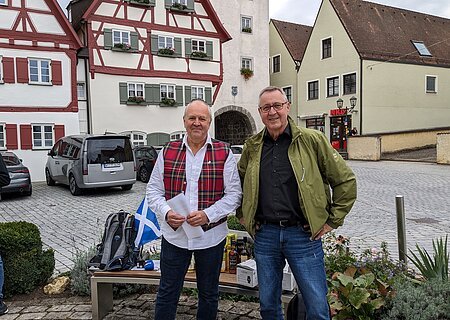 Monheimer Whisky-Tour Stadtführer Rainer Keßler & Whisky-Liebhaber H.P. Schenk Zwei Männer stehen vor Fachwerkhäusern auf gepflastertem Platz und einer Bank mit Rucksack und Flaschen im Hintergrund. Die Bank ist umgeben von Pflanzen. Eine schottische Fahne - in weiß-blau - liegt auf der Sitzbank.