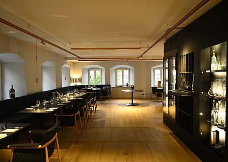 Restaurant im Hotel by Ferber - Hotel, Steakhouse, Bar in der historischen Altstadt von Monheim Innenansicht des Restaurants im Hotel by Ferber, in Monheim. Auf der rechten Seite ein großes schwarzes Regal mit verschiedenen Glasbehältern, Weingläsern und Flaschen. Auf der linken Seite schwarze Holztische mit schwarzen Sitzbänken und schwarzen Stühlen zu sehen.