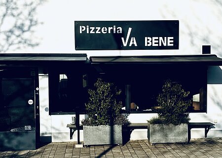 Pizzeria Va Bene - Außenansicht der Pizzeria Außenansicht einer Pizzeria mit schwarzem Schild „Pizzeria VA BENE“, eine Eingansbereich aus Glas, verdunkeltem großen Fenster mit Rollläden und zwei großen Pflanzkübeln davor. Vor der Pizzeria ein gepflasterter Gehweg.