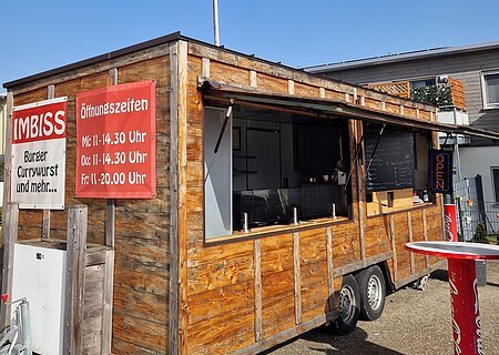 Imbisswagen "Zum dicken Bär" an seinem Stammplatz Holz-Imbisswagen mit geöffnetem Fenster, roten Schildern mit Speisen und Öffnungszeiten, stehend auf Asphalt bei blauem Himmel und einem weiteren Haus im Hintergrund.