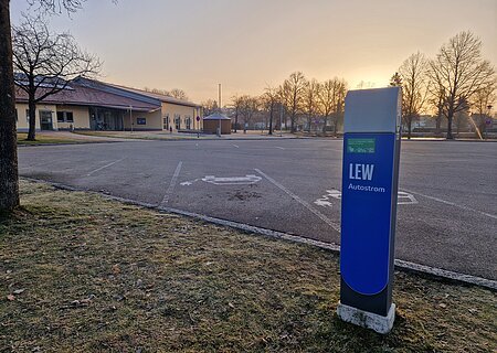 Ladesäule für E-Autos an der Stadthalle Monheim Blaue Ladestation für Elektroautos mit der Aufschrift LEW, auf einem leeren gepflastertem Parkplatz vor einem orangem Gebäude im Hintergrund bei Sonnenaufgang.