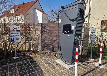 Öffentlich zugängliche Ladesäule an der Raiffeisen-Volksbank Monheim Schwarze Elektroauto-Ladestation mit zwei Ladepunkten stehend vor einem Parkplatz mit Schildern zur Parkzeitbegrenzung auf 2 Stunden, bei Tageslicht. Im Hintergrund Wohnhäuser vor blauem Himmel.