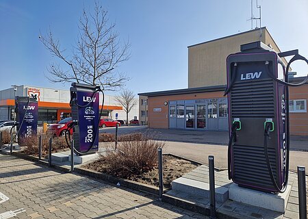 Neue Ladesäulen für E-Autos am EDEKA-Parkplatz in Monheim Drei dunkle Ladestationen für Elektroautos an Parkplätzen mit jeweils zwei Ladepunkten. Im Hintergrund ein oranges Gebäude, der Drogeriemarkt Müller und mehrere parkende Autos daneben.