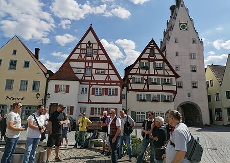 Monheimer Whisky-Touren 2022 Gruppe von Menschen steht auf einem gepflasterten Platz, der Monheimer Innenstadt, vor Fachwerkhäusern und einem Turm mit Uhr. Auf einem gelben Haus davon steht der Schriftzug Nest.