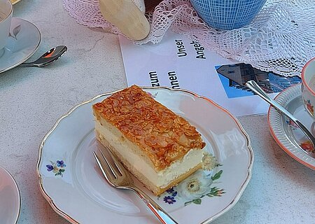 Ehrenamts-Cafè - "Cafè Brunnen" am Seniorenheim Monheim Gedeckter Tisch mit einem Stück Bienenstich-Torte auf dem Teller und einer Gabel im Vordergrund. Außerdem am Tisch in der Mitte des Bildes eine Porzellankanne mit Blumen, daneben eine Puppe mit rosa Schal, gefülltes Wasserglas, umgeben von weiteren Tellern mit Torten. Im Hintergrund der Oberkörper einer Person zu sehen.